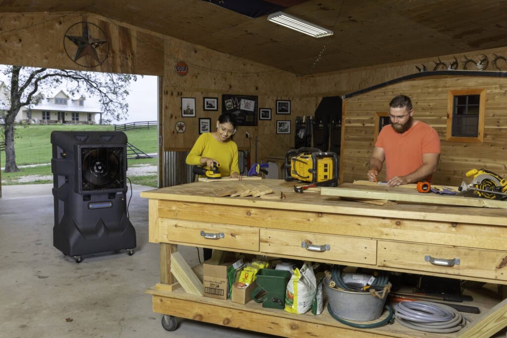 A woman sands wood and a man saws in a spacious workshop. Open windows reveal a rural landscape. Tools and workbench are visible, aiding heat stress prevention.