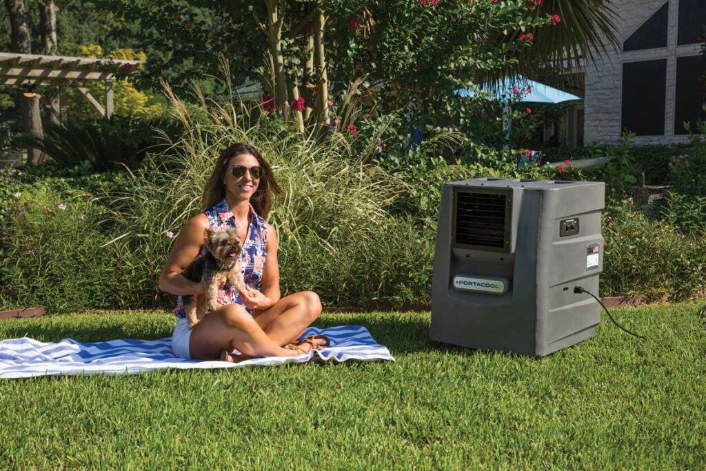 Woman in sunglasses and floral top holding a dog on a towel next to a small Portacool. Scenic background with trees, flowers, and large house.