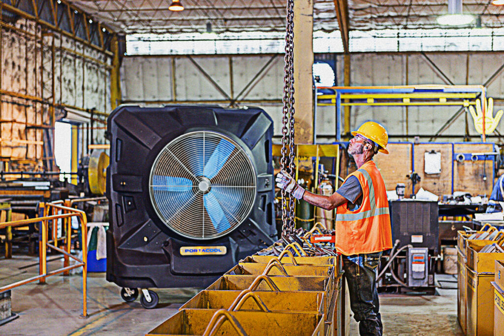 A worker in an orange vest and yellow hard hat expertly operates a chain hoist in a busy industrial warehouse.