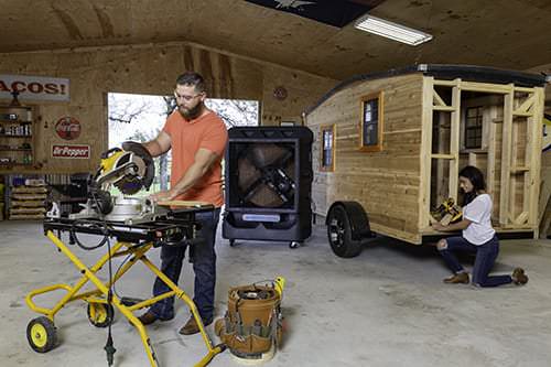 In a workshop, a man uses a miter saw while a woman works beside a wooden trailer. A Cyclone provides comfort amid the equipment.