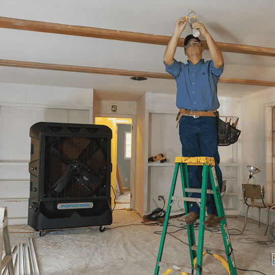 A person in a blue shirt and jeans on a ladder adjusts a light. A Portacool Cyclone 160 is nearby with tools, capturing an HVAC work setting.