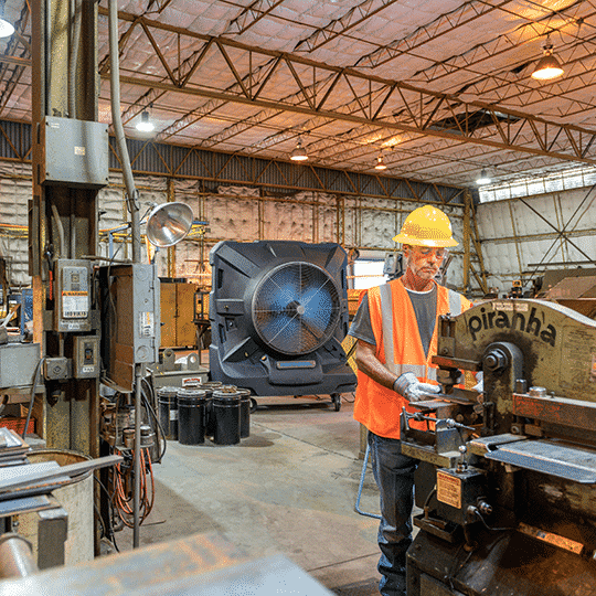 Worker in hard hat and vest operates metal machine in lit warehouse with large tools. Portacool Jetstream 260 fan visible in background.