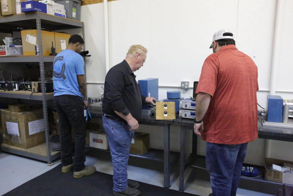 Three people in a workshop: one organizes equipment on a shelf, while the other two examine a device surrounded by electronics. Shelves line the wall.