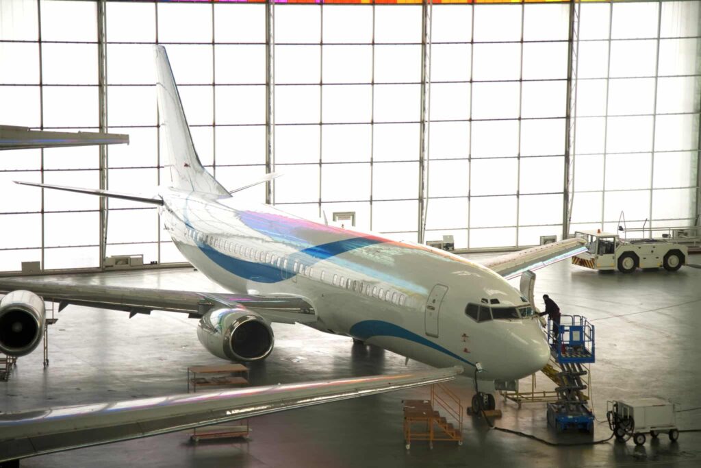 A white and blue airplane in a sunlit hangar. A technician inspects the nose on a platform; nearby are equipment and another plane's wing.