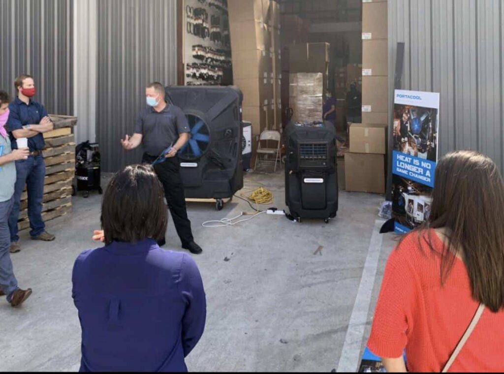 A group listens to a speaker outside a warehouse with large Portacool units and boxes. A "PORTACOOL" sign emphasizes heat management, while some wear masks.