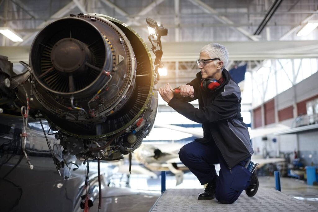 A woman with gray hair and safety glasses kneels on a platform, precisely working on an aircraft engine inside a hangar.