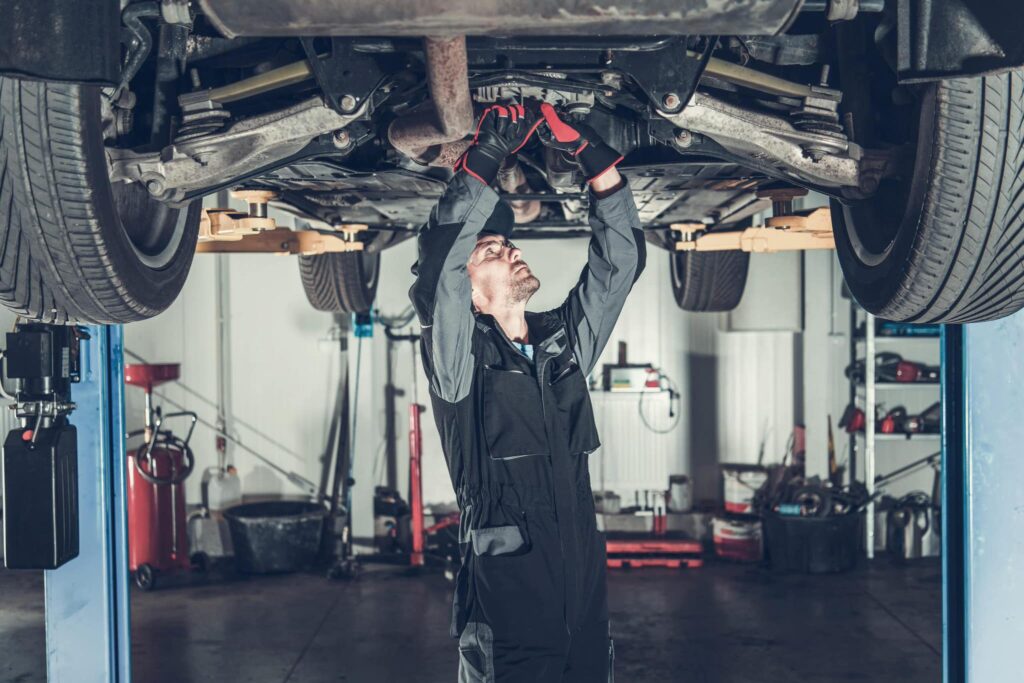 Auto tech in black coveralls works under a lifted car, using tools to ensure precise and effective repairs amidst shop equipment.