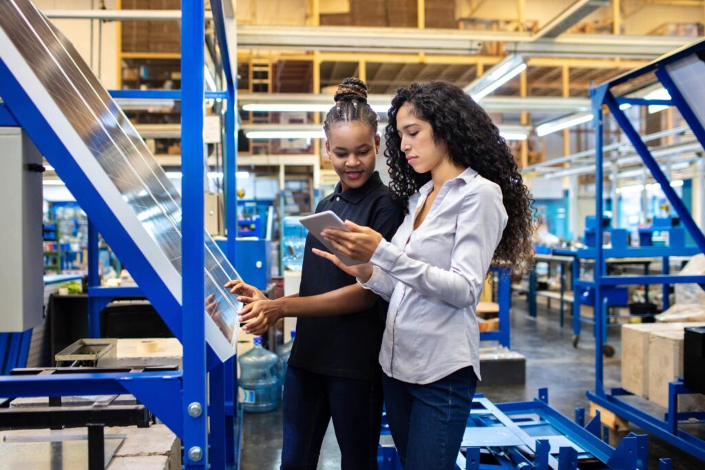 Two women work in a factory; one uses a machine while the other checks a tablet. The bright, spacious area is filled with industrial equipment.