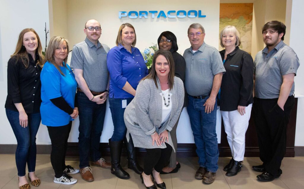 A group of nine casually dressed people pose smiling under a "PORTACOOL" sign in an office lobby.