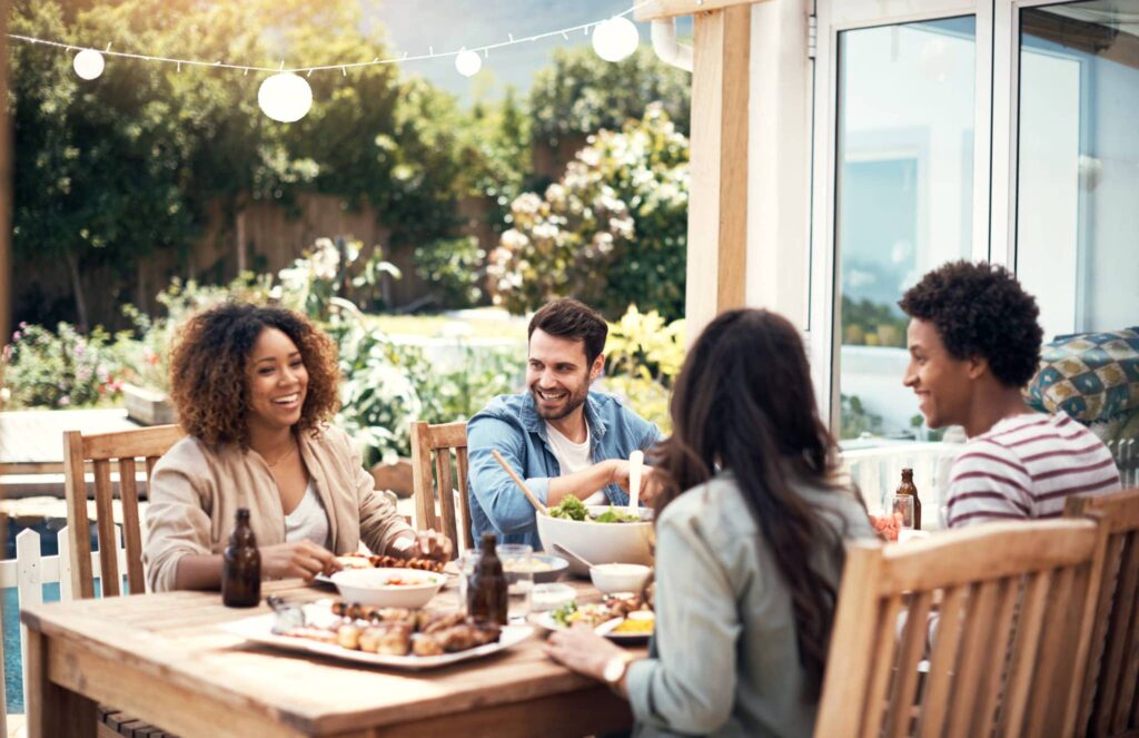 Four friends enjoy a meal at a wooden patio table with food, drinks, and twinkling string lights above. They're surrounded by lush greenery and appear relaxed.