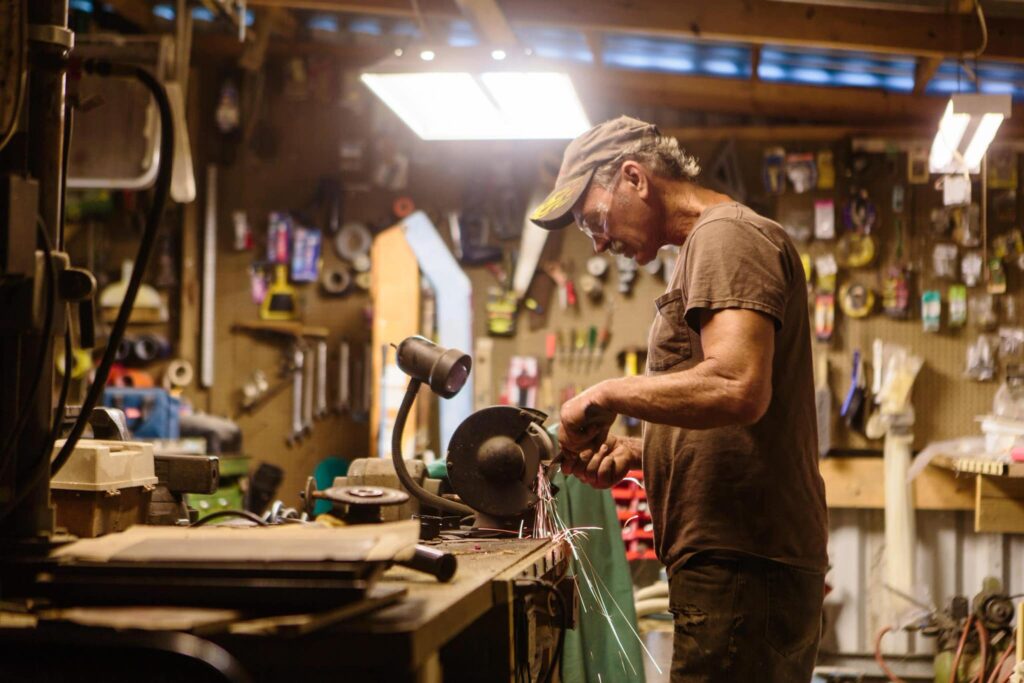 Person in cap and glasses working on metal at cluttered workbench, surrounded by tools. Sparks flying from their precise task.