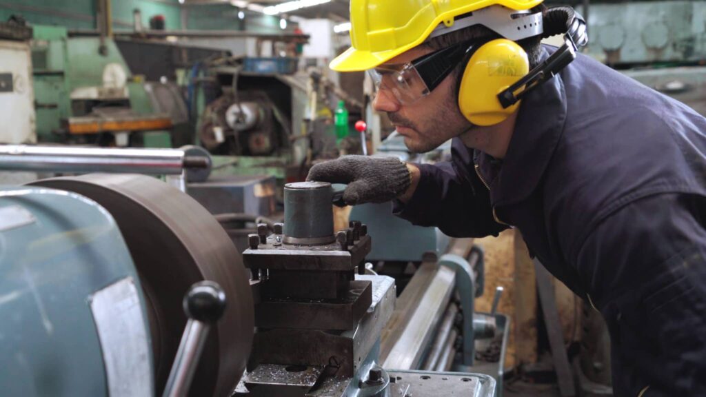 A worker in a yellow hard hat, safety glasses, and ear protection adjusts a lathe component with gloved hands to combat heat stress.