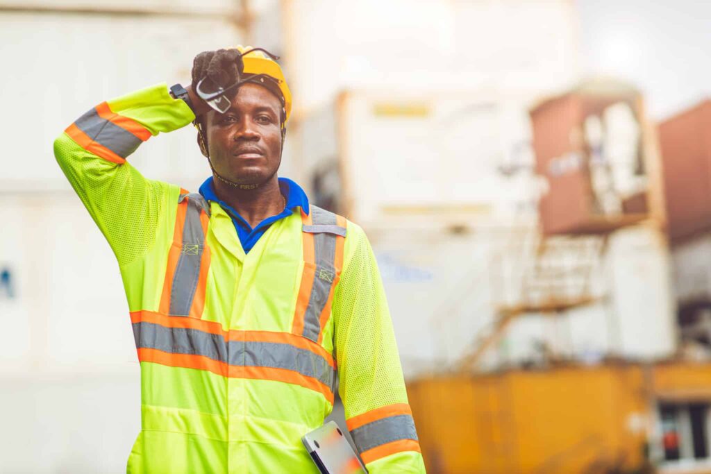 A construction worker in high-visibility gear wipes his forehead, mindful of safety, with blurred shipping containers in the background.