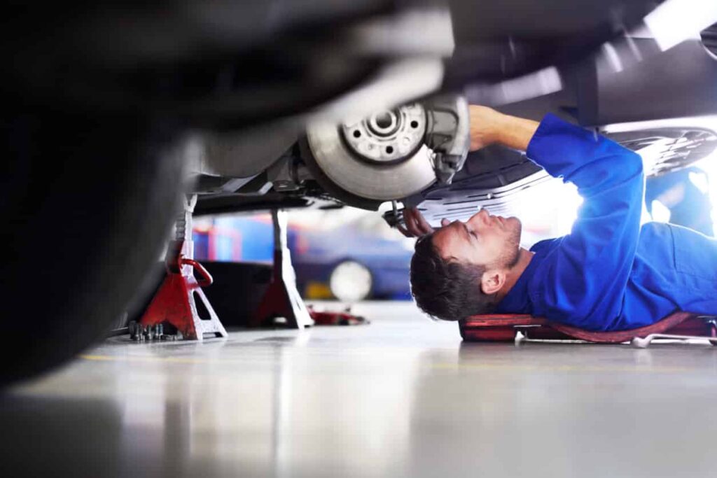 Mechanic in blue jumpsuit on a creeper working under a car elevated on red jack stands. Auto shop showcases expertise and efficient cooling solutions.