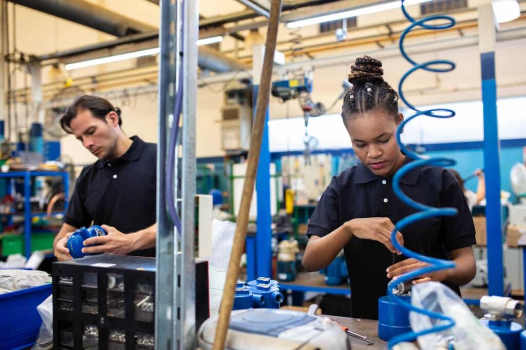 Two workers in black shirts assemble machinery with blue tubing overhead, surrounded by tools. They're focused on heatstroke prevention for safety.