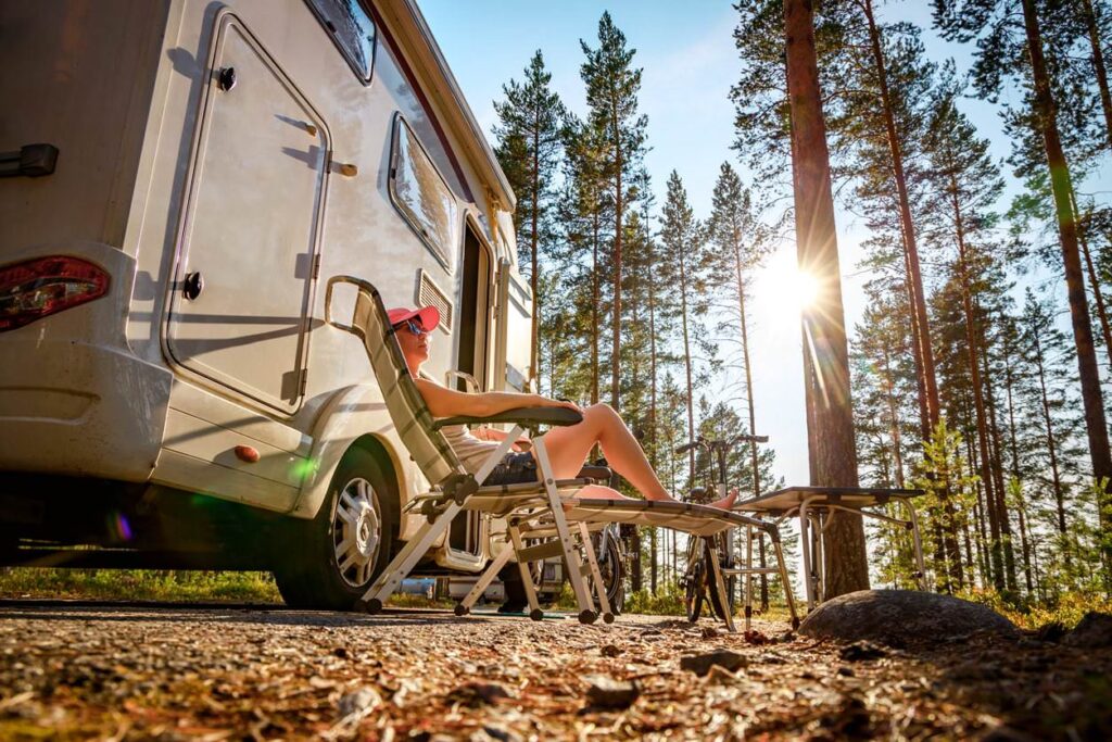 Person relaxing on a sun lounger by RV in forest, wearing cap and sunglasses, with sunlight filtering through tall trees. Scene is peaceful.