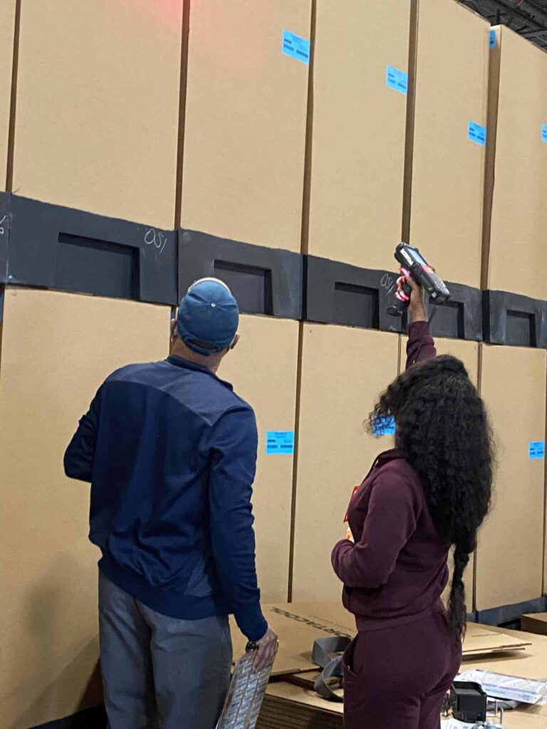 Two people in casual clothing inspect stacked cardboard boxes in a busy warehouse; one scans barcodes, the other notes on a clipboard.