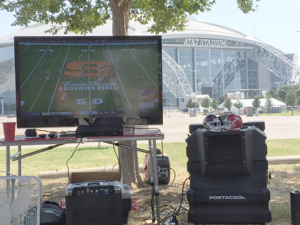 Outdoor setup with a TV playing football, chairs under a tree with helmets, and AT&T Stadium in the background; perfect for cool tailgating.