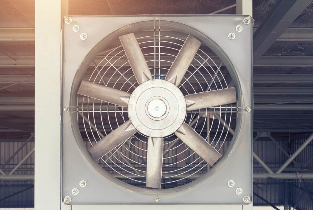 Close-up of a wall-mounted industrial ventilation system with metal blades in a square casing. Sunlight and overhead beams enhance the cooling setup.