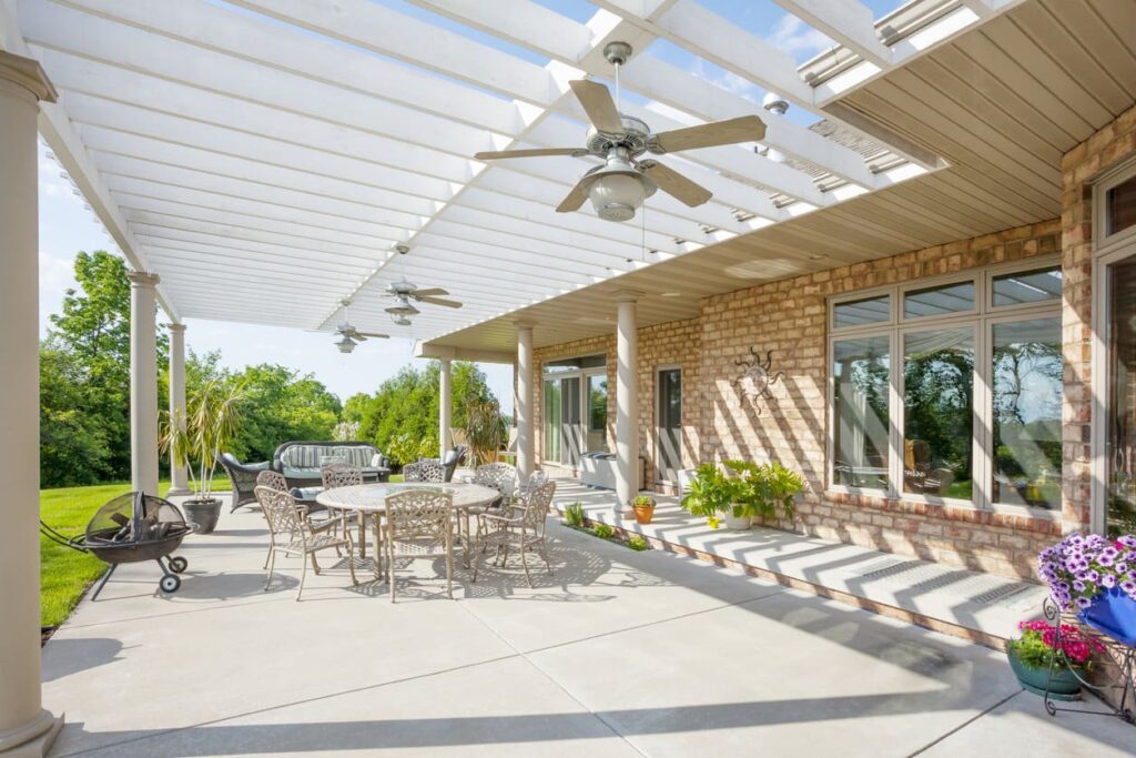 Patio with pergola roof features a round table, chairs, vibrant plants, and lounge area. Cyclone cools the space; lush lawn and clear sky complete the view.