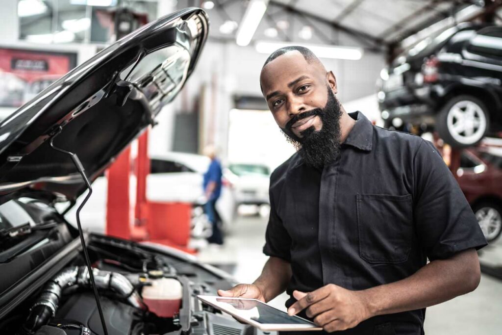 Mechanic with beard holds tablet next to open car hood in busy garage; cars on lifts and someone working in background.