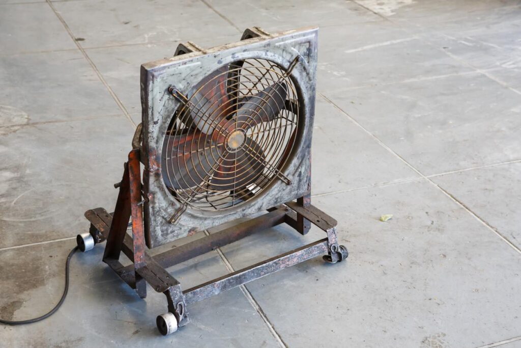 Industrial fan on wheels with metal frame and grill; electrical cord trails from it.