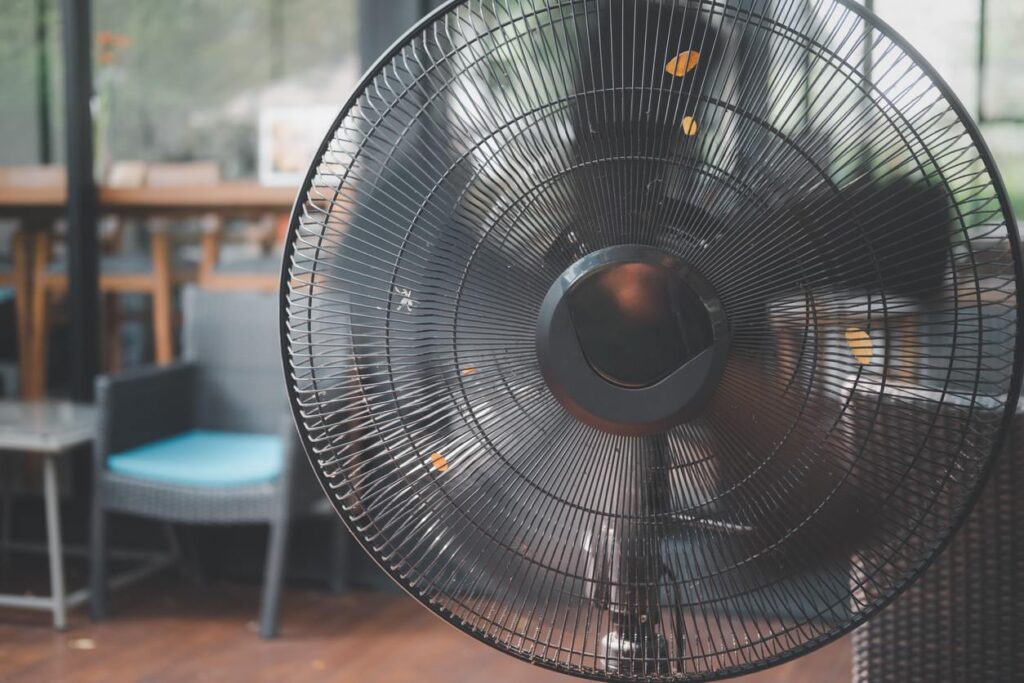 Close-up of a circular fan grille holding orange leaves, set indoors with wicker chairs, table, and sunlit windows.