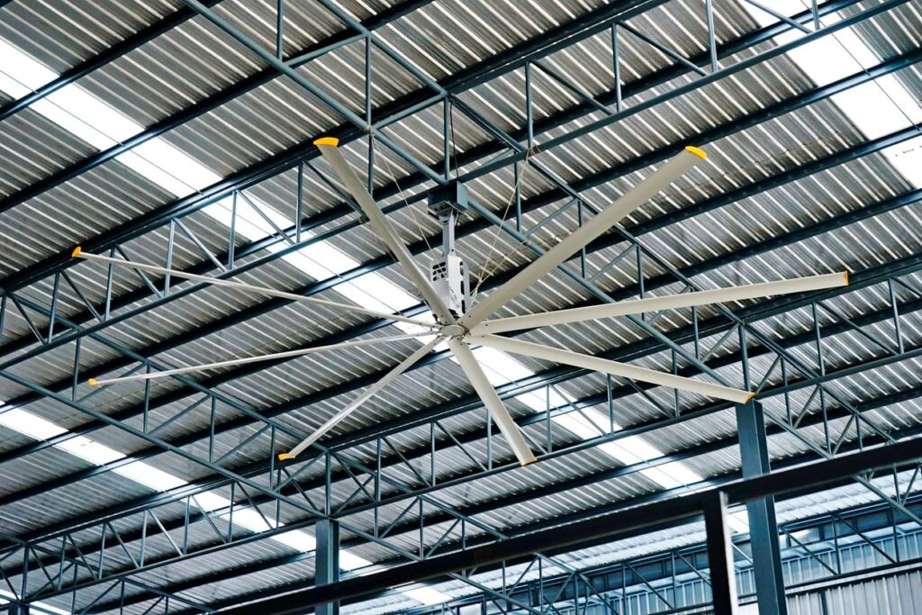 Industrial ceiling fan with multiple blades, yellow tips, hangs in a metal-paneled warehouse. Structural beams visible.