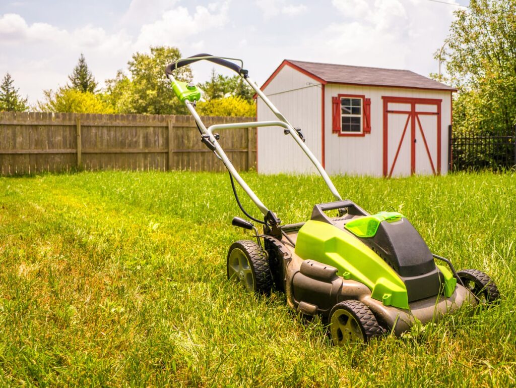 A green lawnmower on a freshly cut lawn. White shed with red trim and a wooden fence in the background, under a partly cloudy sky.