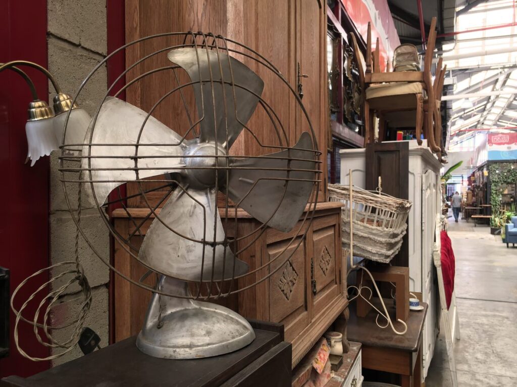 A vintage metal piece on a wooden cabinet in a rustic store aisle, surrounded by antique furniture, an aged lamp, and a wicker basket.