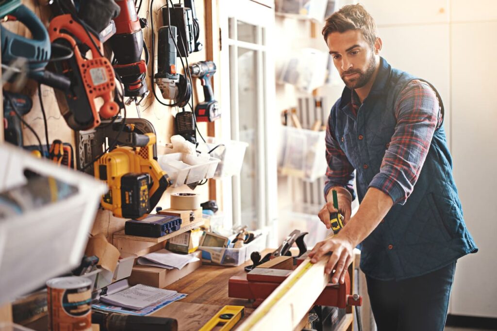 A man in plaid measures wood on a workbench. A swamp cooler maintains comfort in the well-equipped workshop with sunlight streaming through.