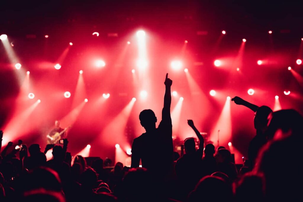 A person raises a finger under bright red stage lights at an indoor concert. A guitarist enhances the vibrant atmosphere in the background.