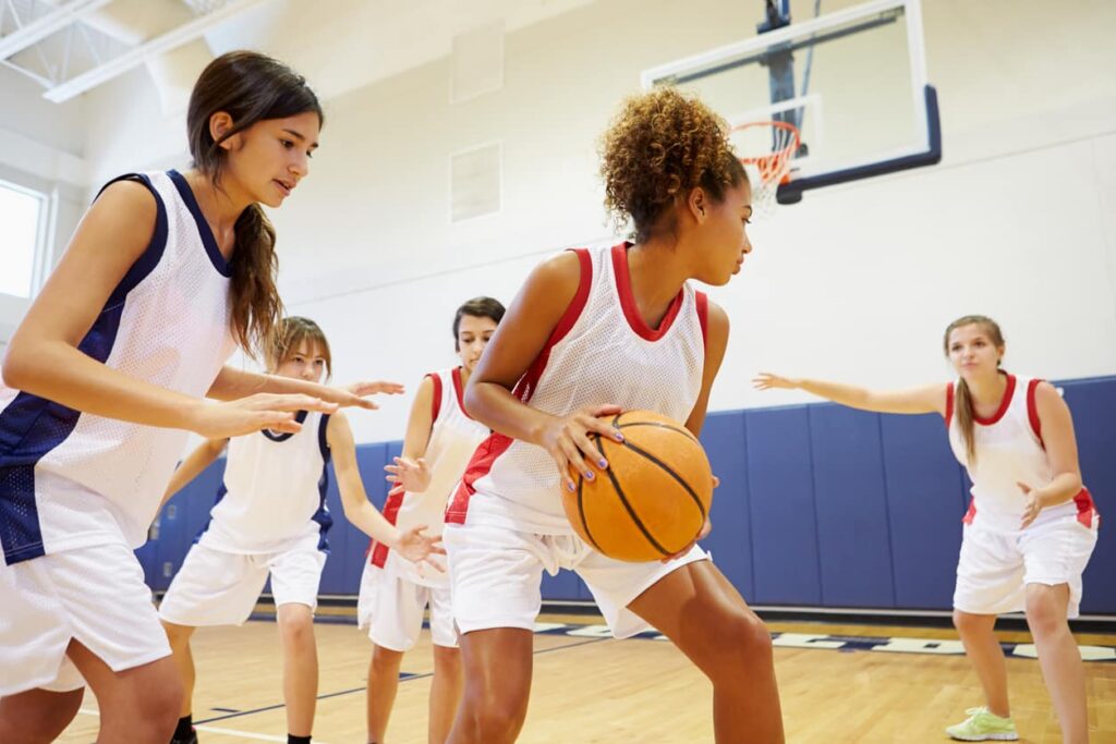 Young basketball players in white and blue jerseys play on a court. One holds the ball, guarded by another, with three nearby. A hoop is in the background.