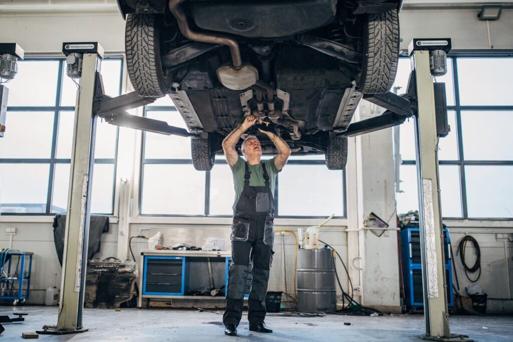 A mechanic inspects a car's underside in a spacious garage with tools and equipment. A cooling system ensures comfort during warm days.