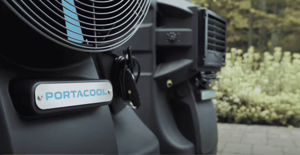 Close-up of a Portacool evaporative cooler with a "PORTACOOL" label on the control panel. Blurred greenery and pavement in the background.