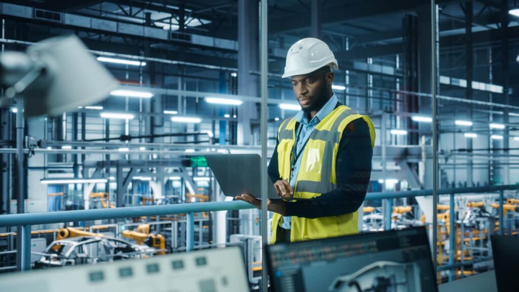 A man in a hard hat and vest uses a laptop in a high-tech factory, with machinery operating in the background.