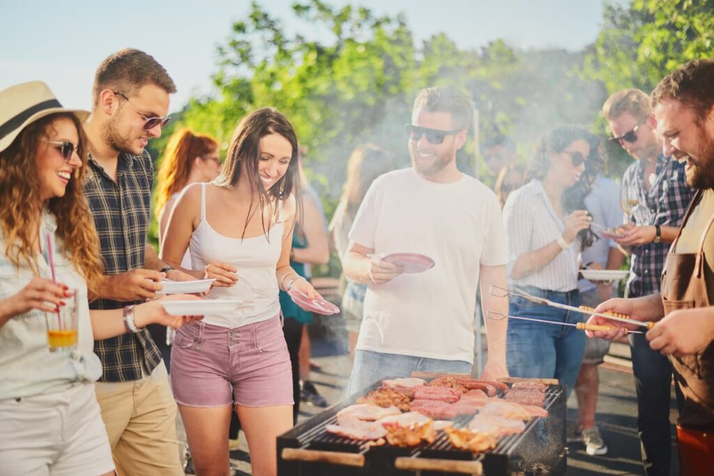 A group enjoys a sunny barbecue on the patio, smiling with plates while meat grills. Misting systems cool the scene, greenery in the background.