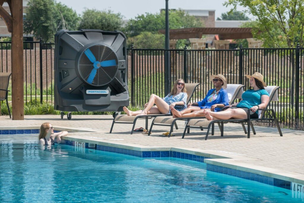 Four people lounge by the pool. One person is in the pool holding onto the side. Trees and a fence create a tranquil setting under a clear sky.