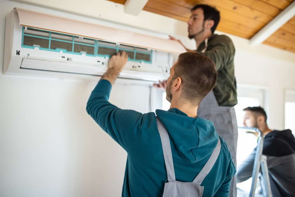 Three individuals install or repair a wall unit in a wood-paneled room. One is on a ladder; two work below. Learn AC installation costs for similar projects.