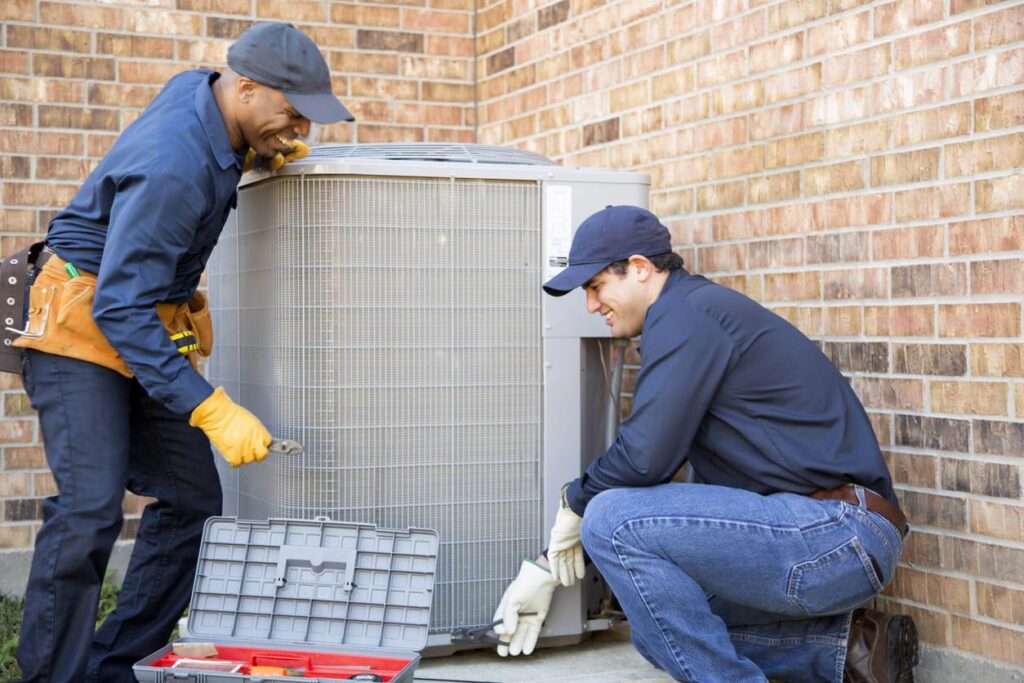 Two technicians in navy uniforms and caps work on an outdoor unit against a brick wall. One kneels with tools, the other stands holding a wrench. An open toolbox is nearby.