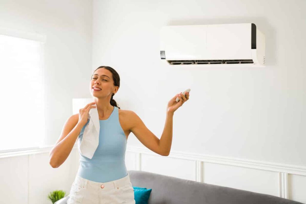 A relieved woman in a blue tank top and white pants holds a remote and towel in her cool, cozy living room.