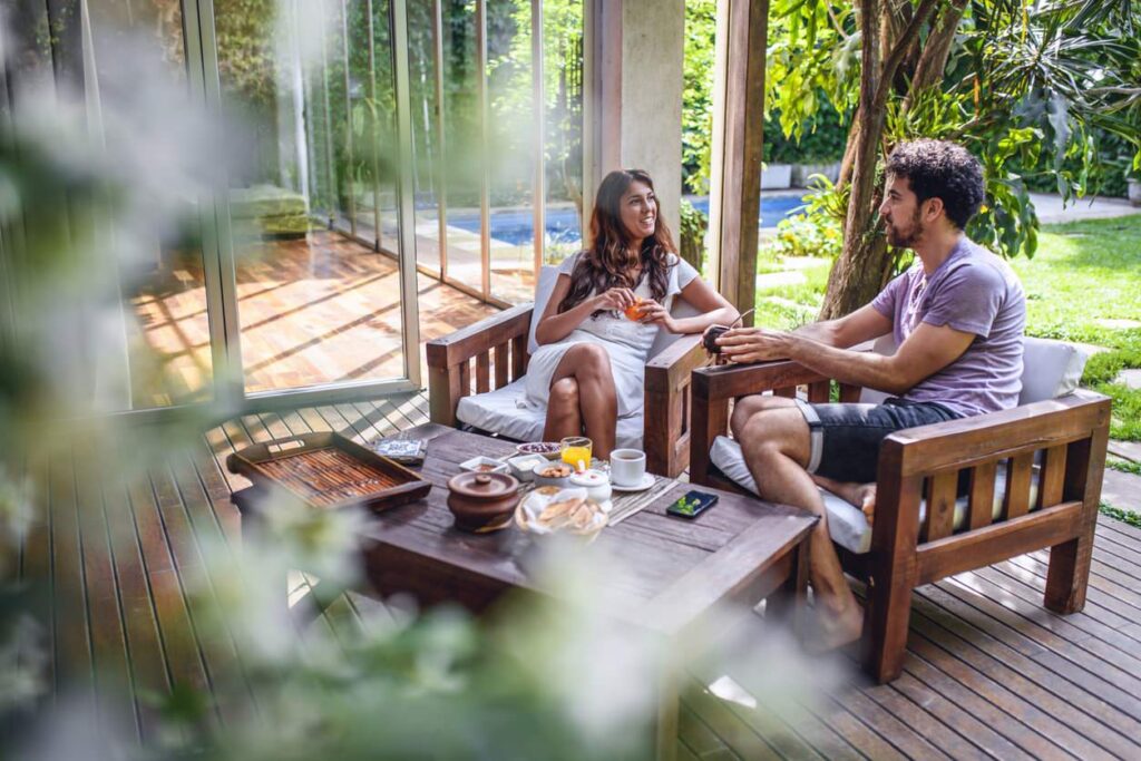 A man and woman sit on wooden chairs with snacks and coffee, conversing in a cozy summer patio surrounded by greenery and large windows.