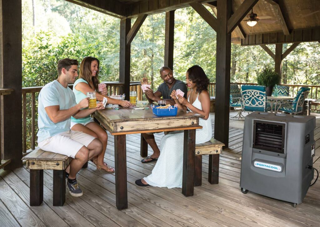 Four people play cards and enjoy drinks at a wooden table on a porch. A Portacool cooler is nearby, with trees in the background.