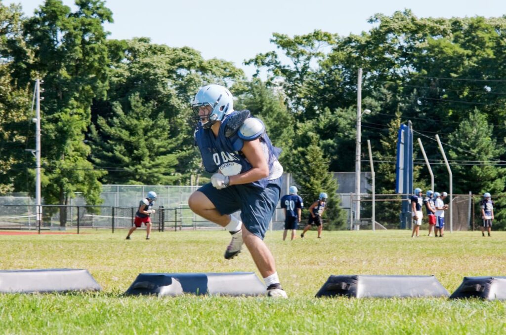 A football player in blue gear practices agility on a sunny field, stepping through pads as teammates and a coach stand nearby, with trees in the background.