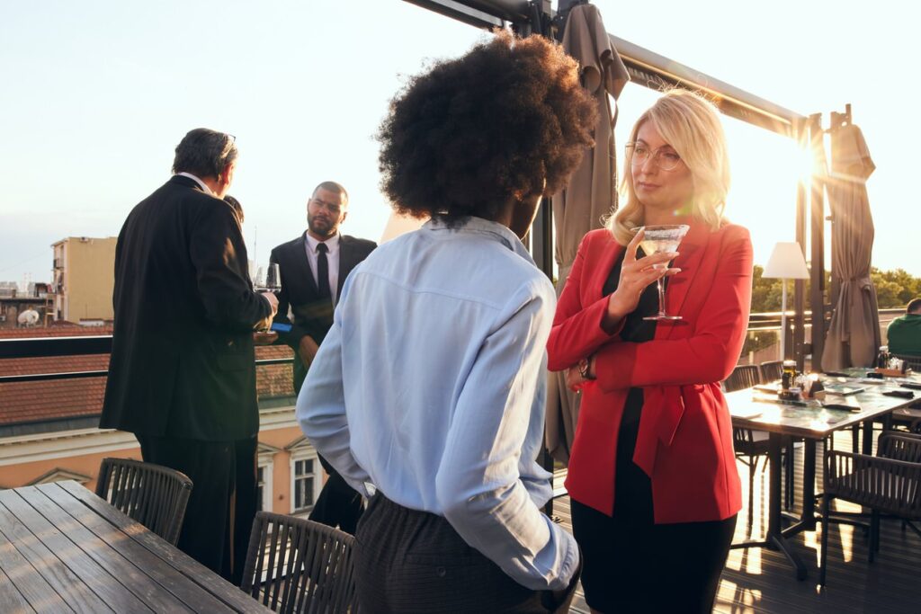 A rooftop summer corporate event at sunset features women chatting over a martini and men in suits against a city skyline.