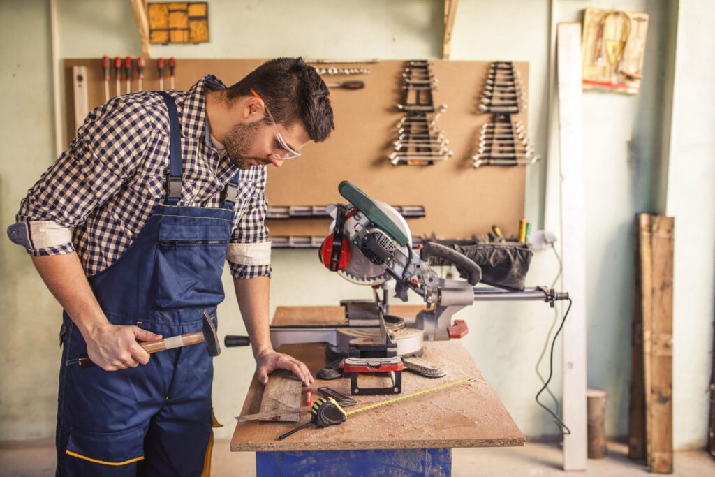 In a cool workshop, a man in checkered shirt and blue overalls uses a chisel near table saw, surrounded by tools; neatly arranged tools on wall behind.