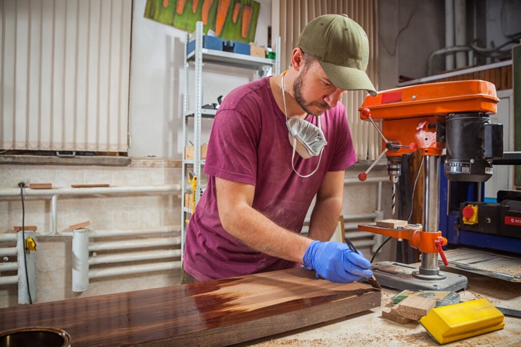 DIY enthusiast in a workshop applies finish to wooden board, surrounded by tools and equipment. They're wearing a cap, face mask, and gloves.