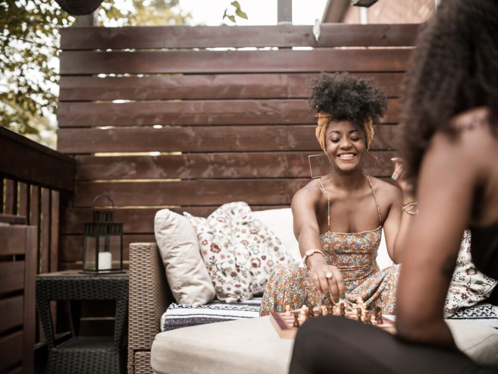 A smiling woman with a headband plays chess on an outdoor patio. She sits on a couch with patterned pillows, surrounded by plants and a wooden fence.