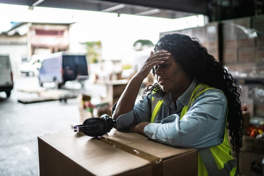 A woman in a yellow safety vest leans on a box in a busy warehouse, looking tired. Headphones are placed beside her.