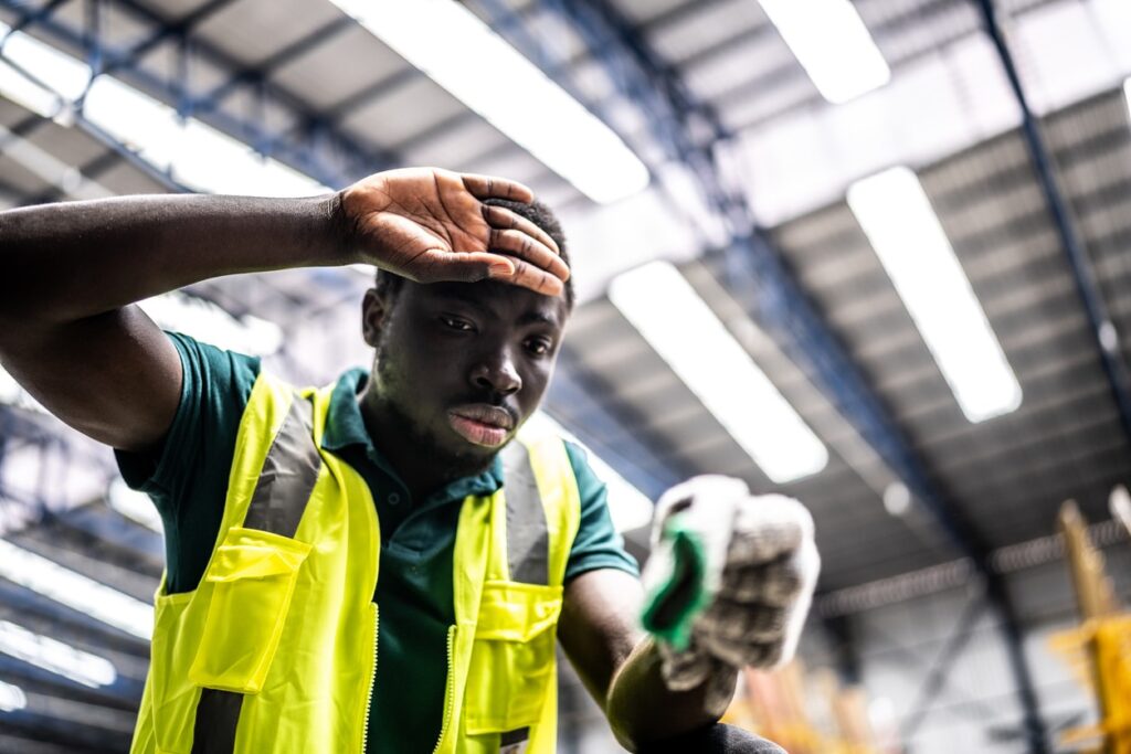 A worker in a yellow safety vest wipes his forehead, tired under bright warehouse lights, yet stays focused on productivity despite the heat.